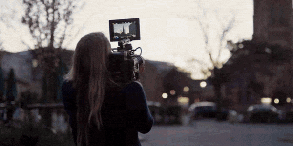 A shot of three dancers on stage mid-synchronized movement; a shot of a woman standing on the countryside looking into a monitor as she angles the camera; a shot of a woman holding a slate surrounded by people in a room mid-camera shot.
